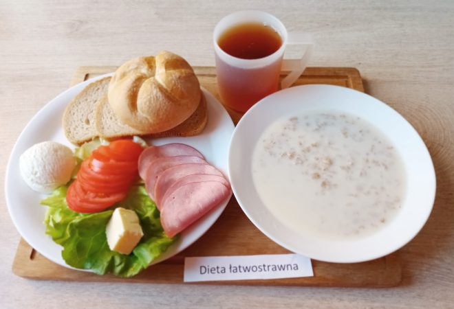 Na talerzu znajduje się śniadanie w diecie łatwostrawnej: plasterki pomidora, wędlina drobiowa, liść sałaty, kostka masła oraz kulka twarożku. Obok ułożono kromki chleba i bułkę. W zestawie znajduje się również kubek herbaty i talerz z mleczną zupą z płatkami. Całość oznaczono etykietą „Dieta łatwostrawna”