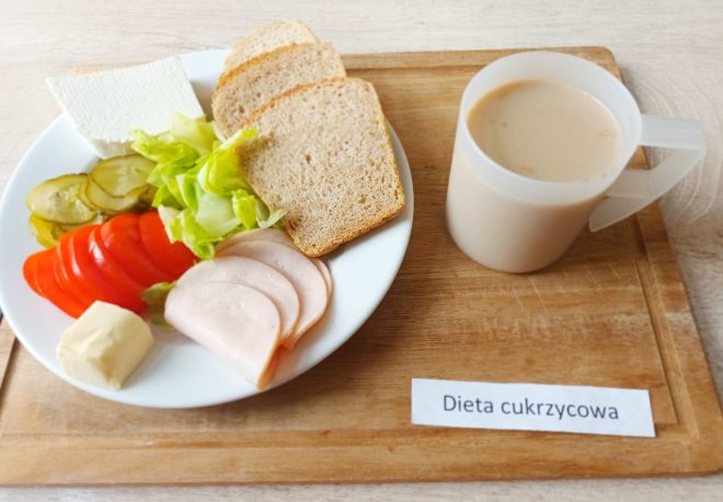 Talerz z trzema kromkami pieczywa, plastrami chudej wędliny, pomidorem, ogórkiem kiszonym, liściem sałaty, porcją twarogu i masłem. Obok stoi kubek jasnego napoju mlecznego. Na tacy znajduje się etykieta „Dieta cukrzycowa”