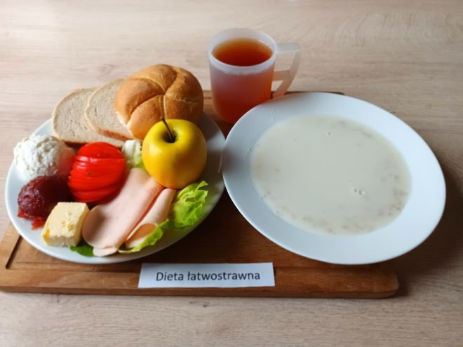 Talerz z dwoma kromkami pieczywa, bułką, plastrami delikatnej wędliny, pomidorem, liściem sałaty, twarożkiem, kawałkiem sera i porcją dżemu. Obok leży jabłko oraz stoi kubek herbaty. W drugim talerzu znajduje się zupa mleczna z kaszą. Zestaw podpisany etykietą „Dieta łatwostrawna”