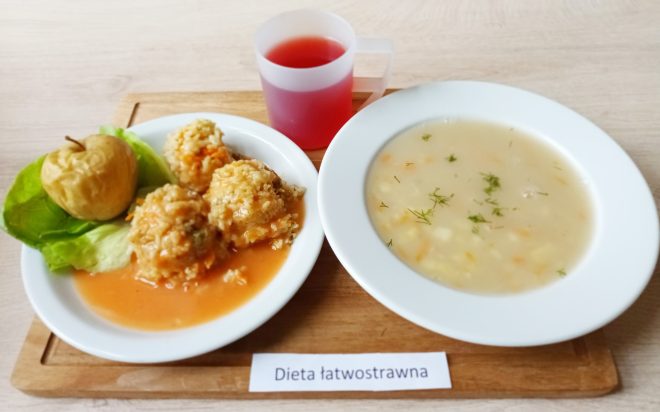 Na talerzu znajdują się pulpety ryżowo-mięsne w jasnym sosie oraz pieczone jabłko ułożone na liściach sałaty. Obok postawiono miseczkę z lekką zupą jarzynową oraz kubek czerwonego napoju. Na tacy widnieje podpis „Dieta łatwostrawna”