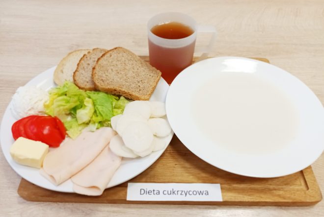 Śniadanie w diecie cukrzycowej: kromki pieczywa pełnoziarnistego, wędlina drobiowa, twarożek, masło, sałata, pomidor oraz plastry białej rzodkwi. Obok herbata bez cukru