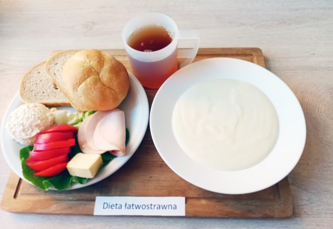 Śniadanie w diecie łatwostrawnej: jasna zupa mleczna w głębokim talerzu oraz talerz z bułką, kromką chleba, twarożkiem, plastrem masła, wędliną drobiową i warzywami. Obok kubek z herbatą. Posiłek podany na drewnianej desce z etykietą „Dieta łatwostrawna”