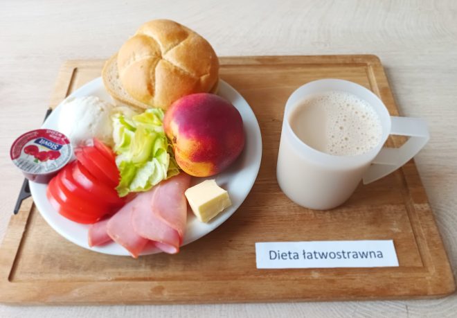 Śniadanie w diecie łatwostrawnej: bułka pszenna, jogurt owocowy, plasterki szynki, masło, sałata i pomidor, brzoskwinia oraz kubek kawy z mlekiem, podane na tacy