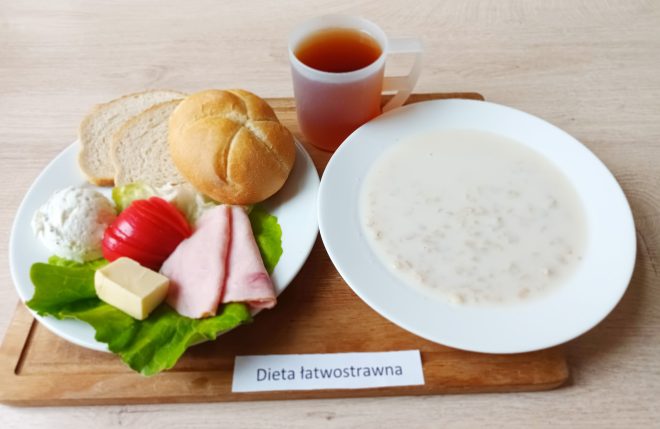 Śniadanie w diecie łatwostrawnej: zupa mleczna z płatkami, pieczywo pszenne, bułka, wędlina drobiowa, twarożek, masło, plaster pomidora, sałata oraz herbata, podane na białych talerzach na drewnianej desce