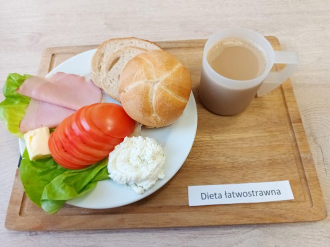 Śniadanie w diecie łatwostrawnej: na białym talerzu bułka pszenna i kromki chleba, plaster szynki drobiowej, ser twarogowy, masło, liście sałaty oraz plasterki pomidora; obok kubek kawy z mlekiem, całość podana na drewnianej tacce