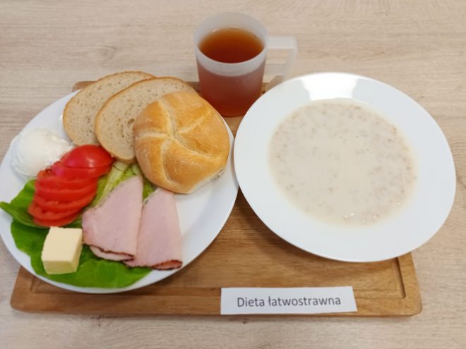 Śniadanie w diecie łatwostrawnej: pieczywo pszenne, bułka, wędlina drobiowa, masło, twaróg oraz warzywa – sałata i pomidor. Obok talerz z zupą mleczną i kubek herbaty, całość na drewnianej desce