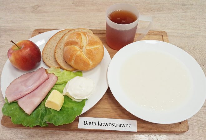 Śniadanie w diecie łatwostrawnej: bułka pszenna i kromki chleba, plasterki szynki, masło, porcja białego twarogu, sałata, jabłko oraz herbata podane na tacy