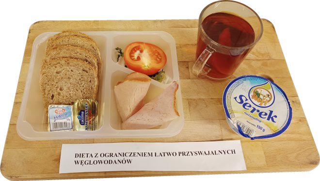 Na plastikowej tacce podzielonej na przegródki znajduje się śniadanie w diecie z ograniczeniem łatwo przyswajalnych węglowodanów: kilka kromek pieczywa pełnoziarnistego, porcja masła i serka topionego, dwa plastry chudej wędliny oraz połówka pomidora na liściach sałaty. Obok tacki stoją szklanka herbaty oraz kubeczek naturalnego serka. Całość ustawiona jest na drewnianej desce, przed którą leży karteczka z nazwą diety