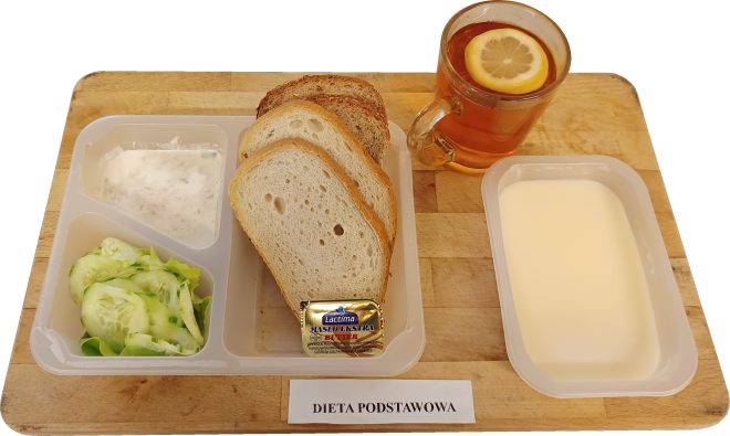 Na drewnianej tacy ułożono zestaw szpitalnego śniadania w plastikowych pojemnikach. W lewym górnym pojemniku znajduje się porcja białego twarożku ze szczypiorkiem. W lewym dolnym pojemniku leży sałatka z plasterków zielonego ogórka. W największym pojemniku pośrodku znajdują się trzy kromki chleba pszennego i razowego oraz porcja masła w złotym opakowaniu. Po prawej stronie ustawiono pojemnik z białą kaszką mleczną. Do posiłku podano szklankę herbaty z plastrem cytryny. Przed tacą widnieje karteczka z napisem „Dieta podstawowa”