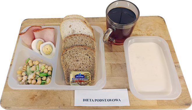 Śniadanie w diecie podstawowej: w pojemniku plastry wędliny, jajko na twardo i sałatka z ciecierzycy, w osobnej przegrodzie trzy kromki pieczywa i masło. Obok kubek herbaty oraz miseczka jasnej zupy mlecznej. Podpis: ‘Dieta podstawowa