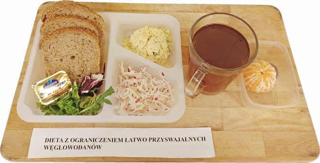 Zestaw śniadaniowy opisany jako Dieta z ograniczeniem łatwo przyswajalnych węglowodanów. W pojemniku znajdują się trzy kromki pieczywa pełnoziarnistego, porcja masła w złotym opakowaniu oraz mała garść miksu sałat. Obok umieszczono pastę jajeczną i surówkę z drobno startego rzodkiewka. Po prawej stronie znajduje się szklanka ciemnego napoju oraz pojemniczek z obranym, podzielonym na cząstki mandarynkiem.