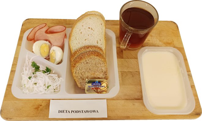 Śniadanie w diecie podstawowej ułożone na drewnianej desce. Na plastikowej tacce znajdują się plastry chudej wędliny, jajko na twardo przekrojone na pół, porcja białej tartej rzodkwi z natką pietruszki oraz kilka kromek pieczywa pszennego i pełnoziarnistego. Obok tacki leży porcja masła, szklanka herbaty oraz oddzielny pojemnik z jasnym jogurtem lub mlekiem. Przed zestawem umieszczono karteczkę z napisem „Dieta podstawowa”