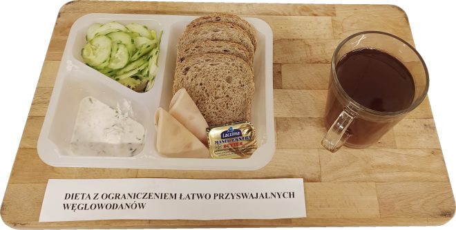 Pojemnik podzielony na trzy części: plasterki ogórka, porcja twarożku z ziołami oraz ciemne pieczywo z dwoma plastrami chudej wędliny i masłem. Obok stoi szklanka herbaty. Etykieta opisuje dietę z ograniczeniem łatwo przyswajalnych węglowodanów