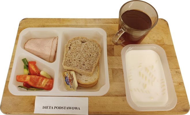 Śniadanie w diecie podstawowej składa się z kilku kromek jasnego i ciemnego pieczywa, porcji masła oraz plastrów szynki. W drugiej przegródce znajduje się pokrojona papryka i ogórek. Po prawej stronie ustawiono kubek herbaty oraz pojemnik z mlekiem i makaronem w kształcie krótkich rurek