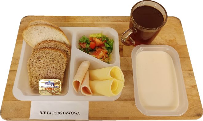 Na tacy znajduje się śniadanie w diecie podstawowej: kilka kromek jasnego i pełnoziarnistego pieczywa, porcja masła, dwa plasterki szynki i dwa plasterki sera żółtego. Obok znajduje się mała porcja sałatki z sałaty, pomidora i ogórka. Po prawej stronie ustawiono kubek herbaty oraz pojemnik z białą zupą mleczną