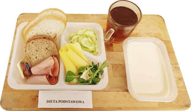 Śniadanie w diecie podstawowej: kromki pieczywa pszennego i razowego, wędlina drobiowa, plasterki sera żółtego, masło, świeży ogórek i mix sałat, podane na tacy z herbatą oraz porcją jogurtu naturalnego