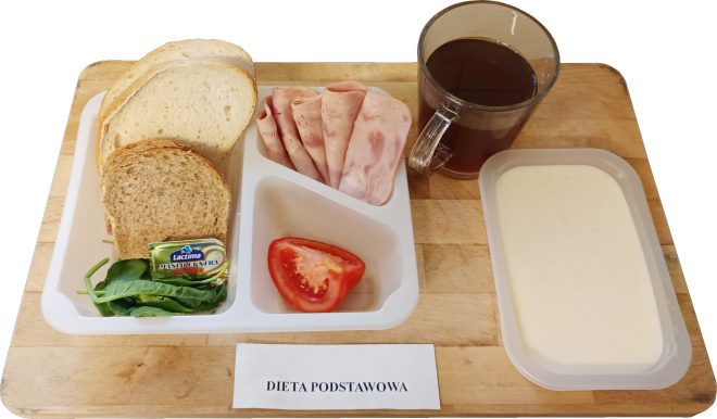 Śniadanie w diecie podstawowej: pieczywo pszenne i razowe, wędlina drobiowa, masło, sałata, pomidor, herbata oraz zupa mleczna, ułożone na tacy na drewnianej desce