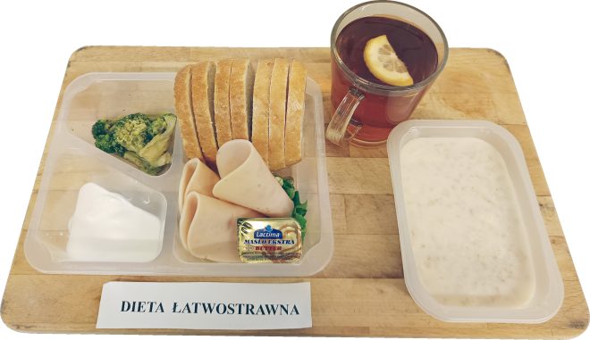 Zestaw śniadaniowy w diecie łatwostrawnej: pieczywo pszenne, wędlina drobiowa, masło, jogurt naturalny, brokuł, herbata z cytryną oraz zupa mleczna, podane w plastikowych pojemnikach na drewnianej desce
