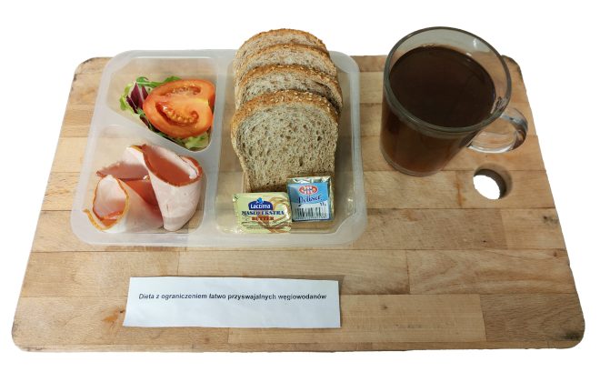 Śniadanie w diecie z ograniczeniem łatwo przyswajalnych węglowodanów: pieczywo pełnoziarniste, wędlina drobiowa, masło, serek topiony oraz warzywa – sałata i pomidor. Zestaw podany w pojemnikach na drewnianej desce, obok kubek herbaty