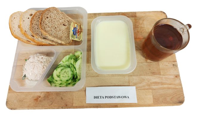 Śniadanie w diecie podstawowej: kromki chleba, porcja masła, pasta twarogowa, świeże plasterki ogórka, zupa mleczna oraz herbata, podane w pojemnikach na drewnianej desce