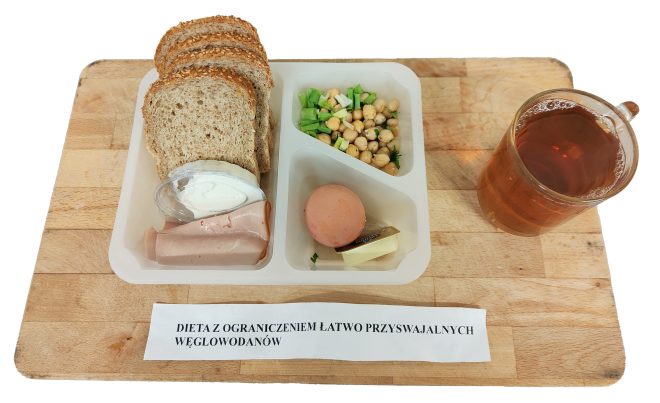 Śniadanie w diecie z ograniczeniem łatwo przyswajalnych węglowodanów: pieczywo pełnoziarniste, wędlina drobiowa, twarożek, sałatka z ciecierzycy i szczypiorku, jajko oraz porcja masła. Obok kubek herbaty. Całość na drewnianej desce z podpisem diety.