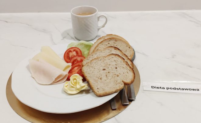 Śniadanie w diecie podstawowej: kromki pieczywa, plasterki wędliny drobiowej, ser żółty, pomidor, sałata i porcja masła, obok kubek napoju
