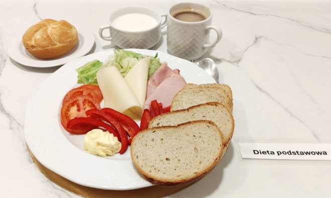 Śniadanie w diecie podstawowej: pieczywo pszenne i żytnie, plasterki sera i wędliny, sałata, pomidor, papryka, masło oraz bułka; w tle kubek mleka i filiżanka kawy