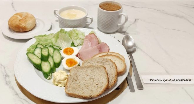 Talerz z jajkami na twardo, plasterkami ogórka, liśćmi sałaty, wędliną, masłem i kromkami pieczywa. Z boku filiżanka kawy, kubek z kaszą manną na mleku i osobno bułka. Podpis: Dieta podstawowa