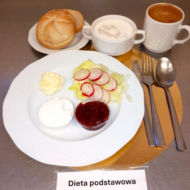Na talerzu ułożono śniadanie: porcję twarożku, marmoladę w formie galaretki, masło oraz sałatę z plasterkami rzodkiewki. Obok znajduje się bułka, kubek z mlekiem z płatkami oraz kubek kawy. Zestaw opatrzono etykietą „Dieta podstawowa”