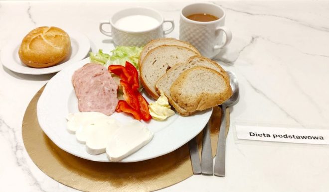 Na talerzu podano śniadanie: kilka kromek chleba, plasterki wędliny, plastry białego sera, liście sałaty i paski czerwonej papryki oraz porcję masła. Obok znajduje się bułka kajzerka, kubek z mlekiem i kubek z herbatą. Zestaw opisany jest etykietą „Dieta podstawowa”