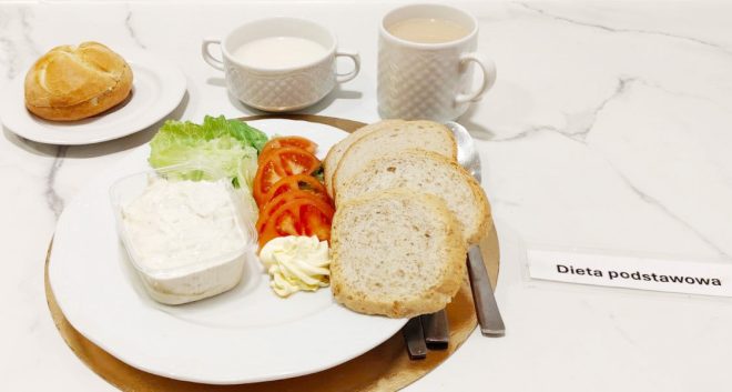 Śniadanie w diecie podstawowej podane na białym talerzu ustawionym na złotej podkładce. Na talerzu znajdują się trzy kromki pieczywa pszennego, plasterki pomidora, liście sałaty, porcja masła oraz miseczka z twarożkiem. W głębi stoją dwie filiżanki: jedna z mlekiem, druga z kawą z mlekiem. Obok na oddzielnym talerzyku leży bułka kajzerka. Po prawej stronie widoczna jest karteczka z napisem „Dieta podstawowa”