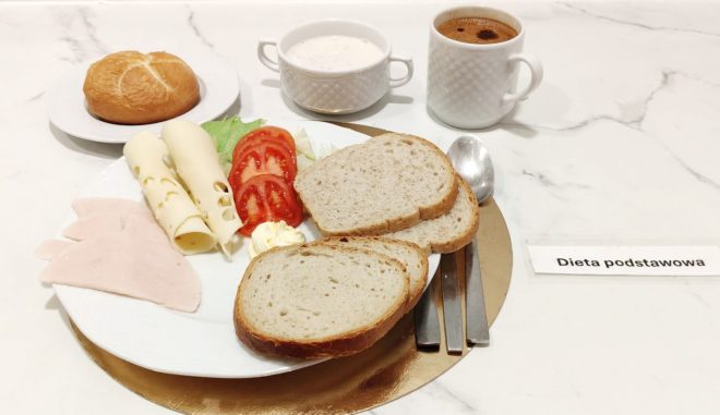 Talerz śniadaniowy z trzema kromkami pieczywa, plastrami wędliny z indyka, zwiniętymi plastrami sera żółtego, masłem, liściem sałaty i plasterkami pomidora. Obok stoi kubek kawy, miseczka jogurtu lub deseru mlecznego oraz oddzielnie podana bułka. Z prawej strony widoczna etykieta „Dieta podstawowa”