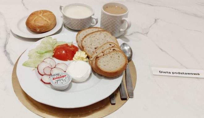 Śniadanie w diecie podstawowej: pieczywo pszenne i mieszane, jogurt naturalny, rzodkiewka, pomidor, sałata, masło oraz napoje mleczny i gorący, podane na talerzu na jasnym blacie