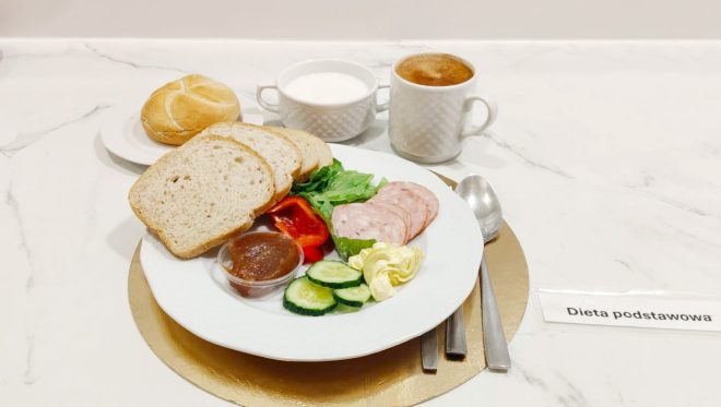 Śniadanie w diecie podstawowej: kromki pieczywa pszennego, wędlina drobiowa, masło, świeże warzywa – ogórek, papryka i sałata – oraz porcja dżemu. Do posiłku kawa i mleko