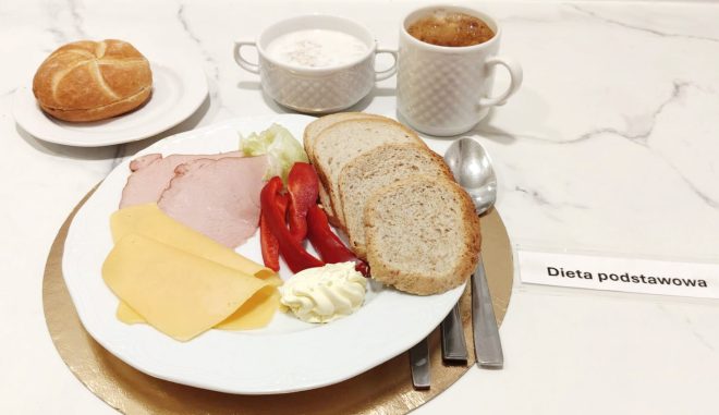 Śniadanie w diecie podstawowej: pieczywo pszenne i razowe, wędlina drobiowa, plasterki sera żółtego, masło oraz świeże warzywa. W tle zupa mleczna i kawa zbożowa
