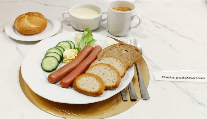 Śniadanie w diecie podstawowej: parówki, kromki pieczywa pszennego i razowego, ogórek świeży, sałata oraz porcja masła. Na stole bułka, miseczka z mlekiem i kubek kawy