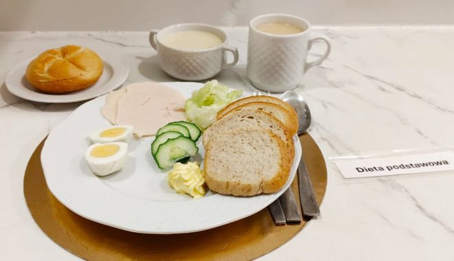 Śniadanie w diecie podstawowej: kromki chleba, jajko na twardo, wędlina drobiowa, ogórek świeży, sałata oraz masło; w tle bułka, zupa mleczna i herbata