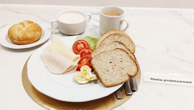 Śniadanie w diecie podstawowej: kromki chleba, wędlina drobiowa, plaster sera, pomidor, sałata oraz masło; w tle bułka, zupa mleczna i herbata