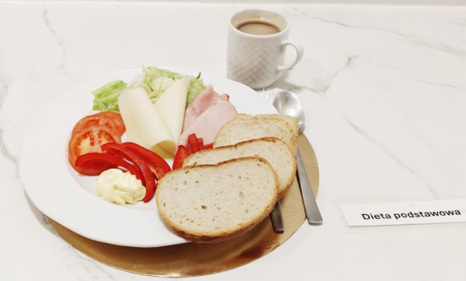 Śniadanie w diecie podstawowej: pieczywo, plasterki sera i wędliny, sałata, pomidor, papryka oraz masło; w tle filiżanka kawy