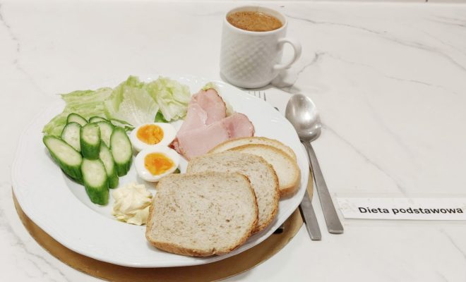 Śniadanie w diecie podstawowej: na talerzu znajdują się kromki pieczywa, jajka na twardo, plasterki świeżego ogórka, liście sałaty, masło oraz plastry wędliny. Obok stoi filiżanka kawy oraz komplet sztućców. Podpis: Dieta podstawowa