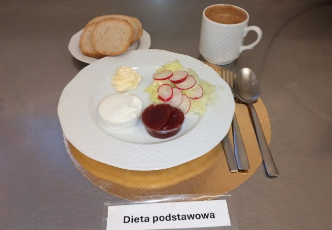 Śniadanie na białym talerzu: twarożek, masło, sałata z plasterkami rzodkiewki oraz porcja dżemu. W tle talerzyk z pieczywem i kubek kawy. Pod talerzem widoczna etykieta ‘Dieta podstawowa