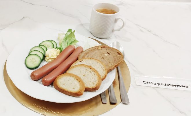 Śniadanie w diecie podstawowej: parówki, kromki pieczywa pszennego i razowego, ogórek świeży, sałata oraz porcja masła. Obok kubek gorącego napoju