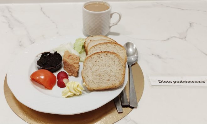 Śniadanie w diecie podstawowej: kromki pieczywa pszennego, pasztet, porcja masła, rzodkiewki, sałata oraz pomidor. Na stole kubek kawy z mlekiem
