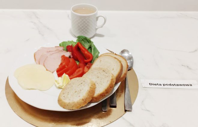 Śniadanie w diecie podstawowej: kromki pieczywa, ser żółty, wędlina, pomidor, papryka i sałata, obok kubek ciepłego napoju