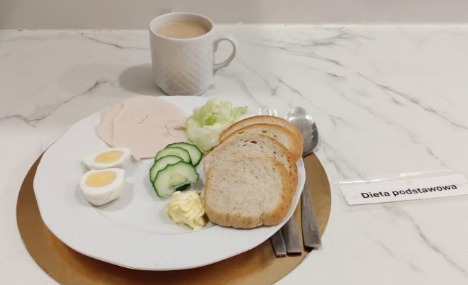 Śniadanie w diecie podstawowej: kromki chleba, jajko na twardo, wędlina drobiowa, ogórek, sałata i masło, obok kubek kawy z mlekiem