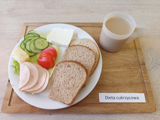 Śniadanie w diecie cukrzycowej: na białym talerzu dwie kromki pieczywa razowego, plasterki wędliny drobiowej, ser biały, masło oraz świeże warzywa – ogórek, sałata i pomidor. Obok kubek z kawą lub herbatą z mlekiem. Całość podana na drewnianej tacy z opisem „Dieta cukrzycowa”