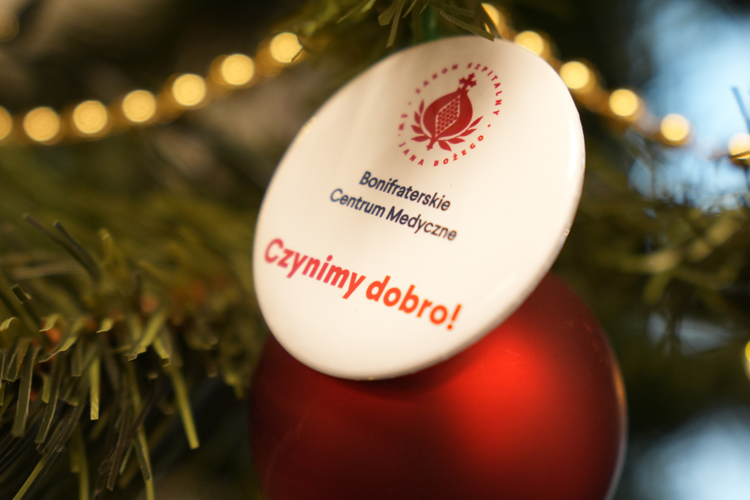 Świąteczna choinka udekorowana czerwoną bombką oraz białą zawieszką z logo Bonifraterskiego Centrum Medycznego i hasłem »Czynimy dobro!«, w tle ciepłe, rozmyte światełka