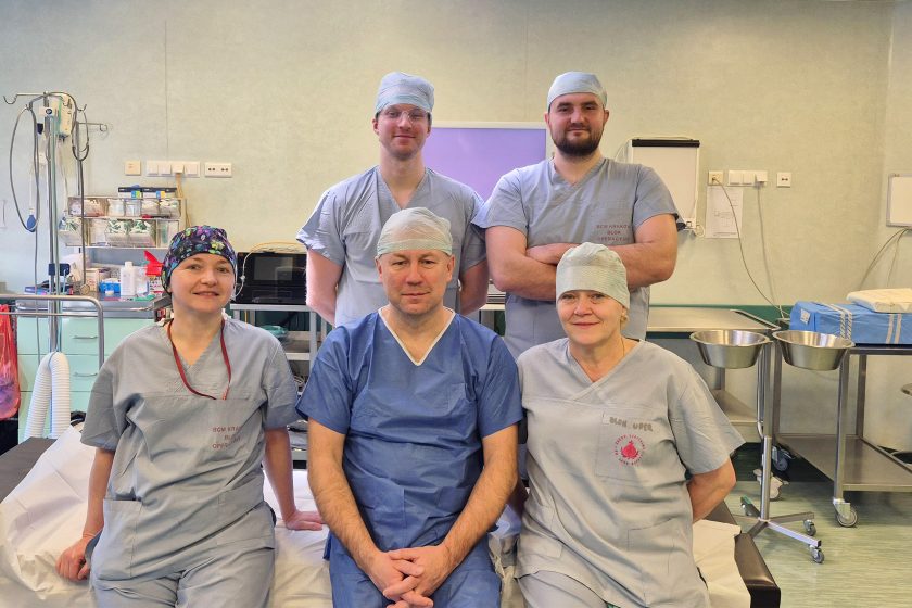 Grupowe zdjęcie zespołu medycznego w sali operacyjnej: sześć osób ubranych w stroje chirurgiczne i czepki. Trzy osoby siedzą na stole zabiegowym, pozostałe stoją za nimi. W tle widoczna aparatura medyczna, monitory, wózki zabiegowe oraz lampa operacyjna. Wszyscy patrzą w stronę aparatu; fotografia ma charakter zespołowy i dokumentacyjny