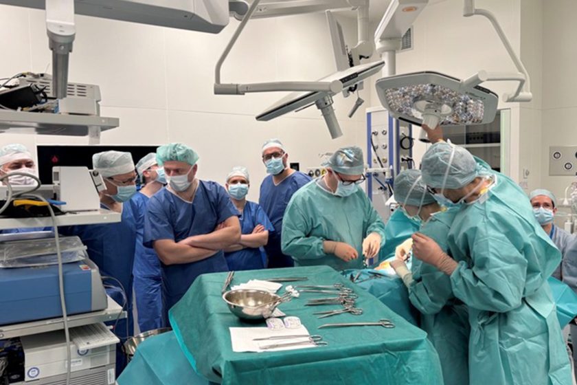 Na sali operacyjnej zespół lekarzy ubranych w sterylne zielone i niebieskie fartuchy oraz maseczki wykonuje zabieg chirurgiczny przy stole operacyjnym. Część uczestników obserwuje procedurę z bliska, stojąc wokół pola operacyjnego. Na stoliku widoczne są narzędzia chirurgiczne. Zdjęcie wykonano podczas warsztatów poświęconych laparoskopowemu leczeniu przepuklin