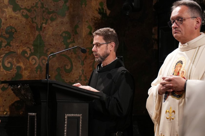 Zakonnik w czarnym habicie czyta tekst przy pulpicie z mikrofonem w kaplicy, obok stoi kapłan w białych szatach liturgicznych. W tle dekoracyjne, historyczne wnętrze